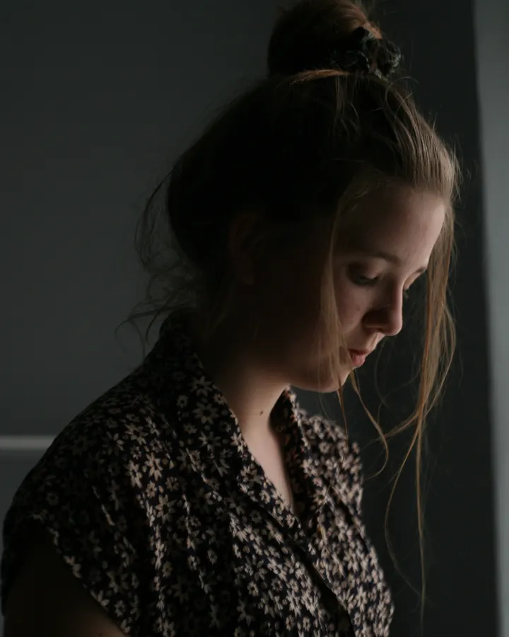 A woman in a floral blouse looking down, lost in thought by a window